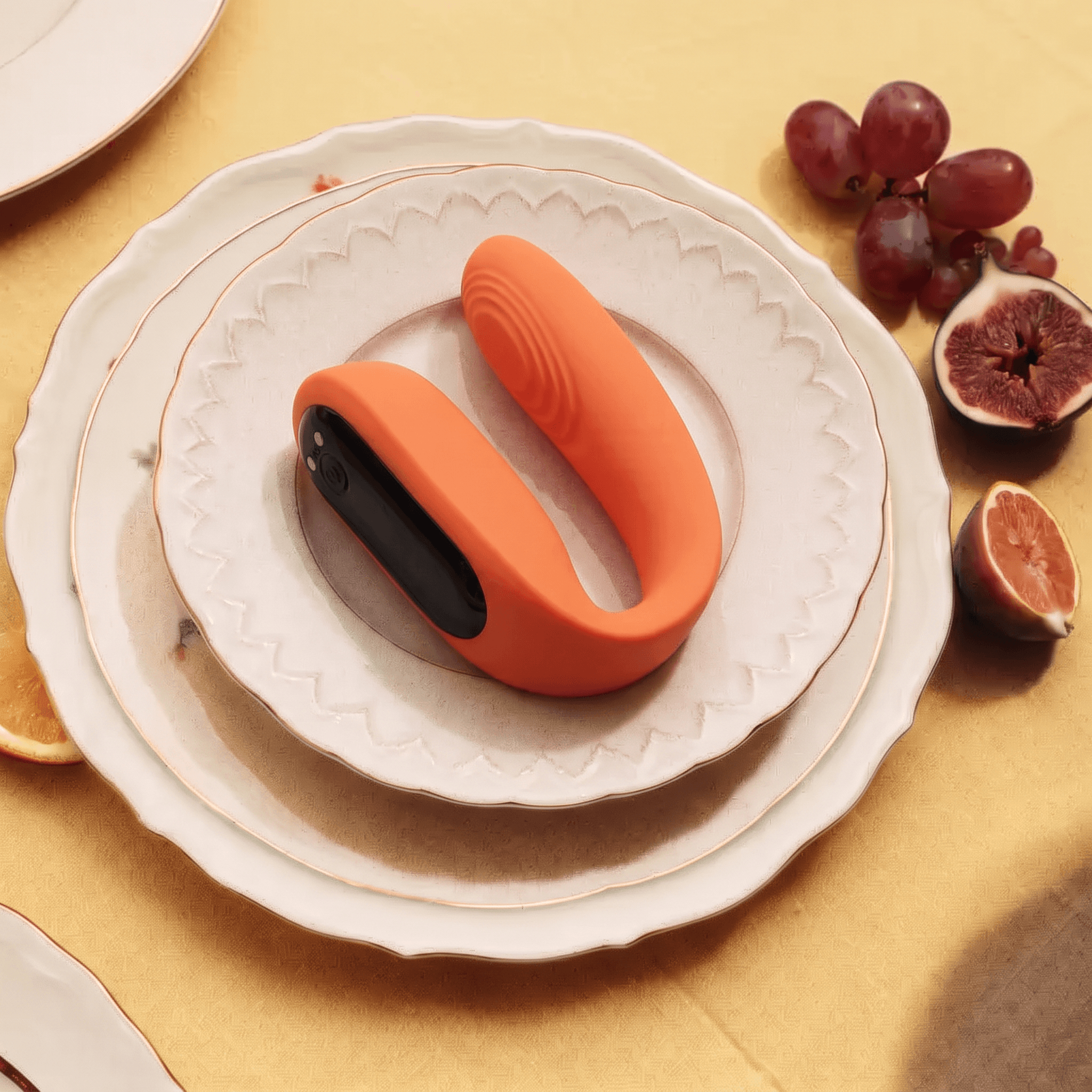 A coral couple vibrator resting on a rimmed plate beside figs and grapes