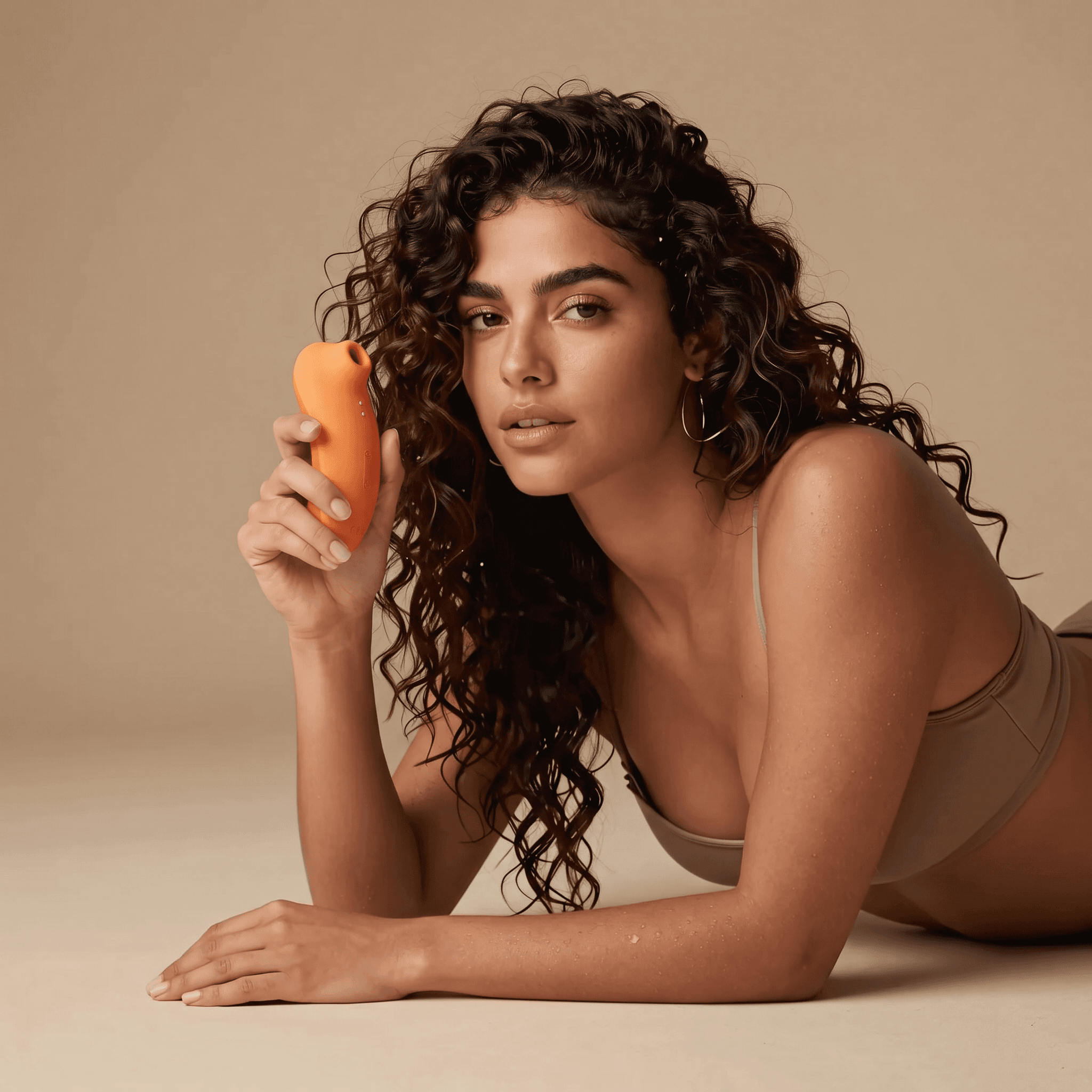 A woman lying on a warm sand backdrop, holding the coral air-pulse sucker beside her face